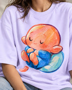 Close-up of a sleeping orange-headed creature curled on a blue cushion printed on a lavender oversized French terry t-shirt