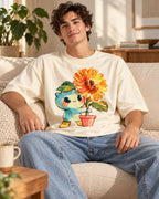 Man on a sofa in an off-white oversized French terry t-shirt with a teal creature carrying a potted orange sunflower
