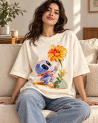 Woman on a sofa in an off-white oversized French terry t-shirt with a blue alien in a red scarf reaching for an orange flower