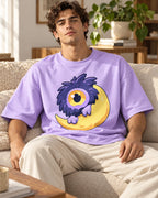 Man on a sofa in a lavender oversized French terry t-shirt with a fluffy one-eyed purple monster on a yellow moon
