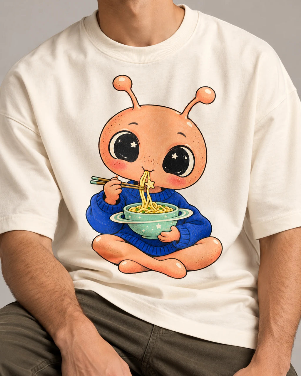 Close-up of a peach alien eating noodles from a ringed bowl printed on an off-white oversized French terry t-shirt
