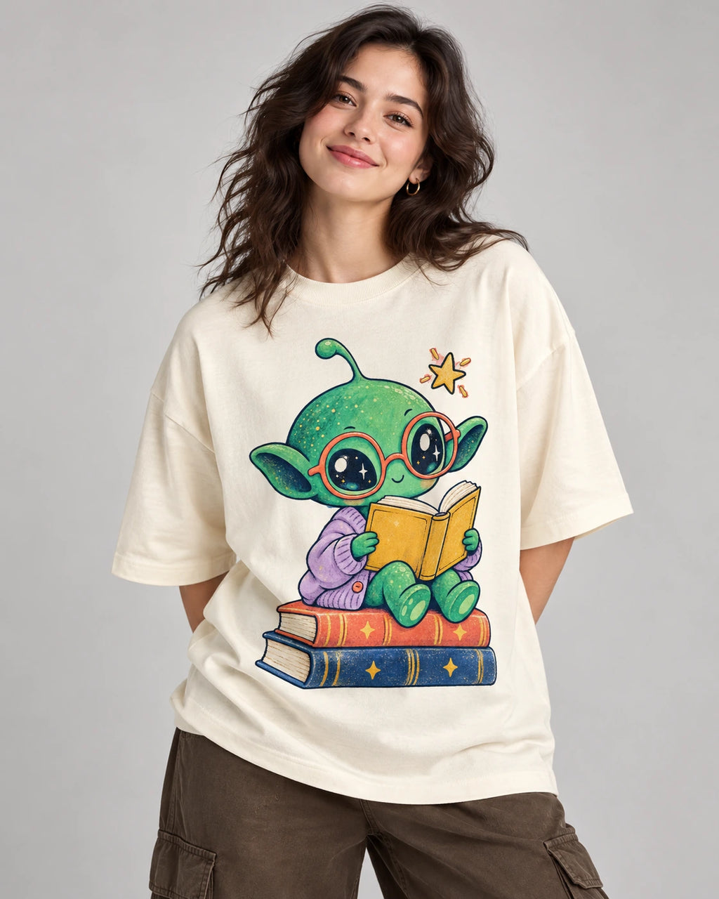 Woman in an off-white oversized French terry t-shirt printed with a green alien in red glasses reading on a stack of books