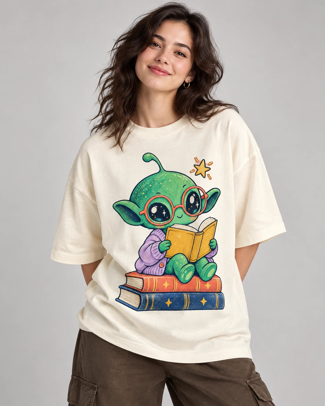 Woman in an off-white oversized French terry t-shirt printed with a green alien in red glasses reading on a stack of books