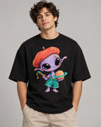 Man in a black oversized French terry t-shirt printed with a purple alien in a beret painting a ringed planet