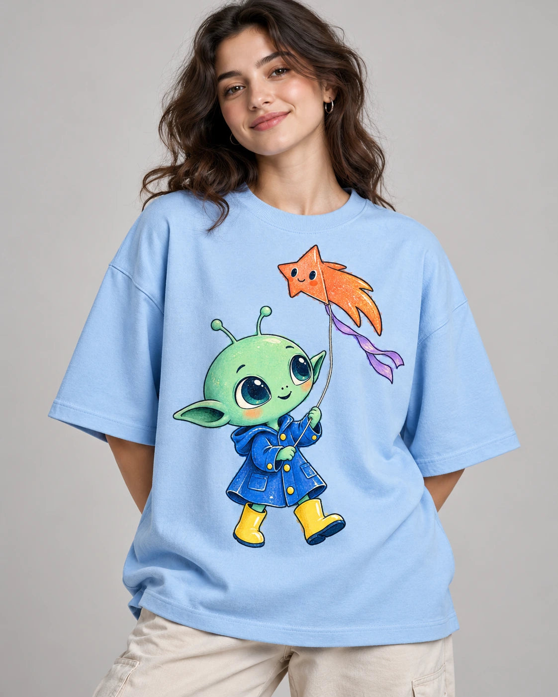 Woman in a baby blue oversized French terry t-shirt printed with a green alien in a raincoat flying a star-shaped kite